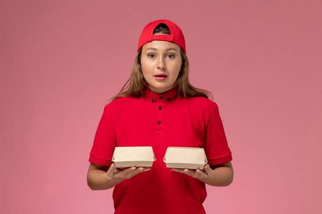 Corriere femminile di vista frontale in uniforme rossa e mantello che tiene i pacchetti di cibo di consegna sulla parete rosa-chiaro, lavoratore di lavoro dell'azienda di servizio di consegna uniforme