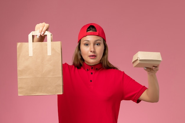 Corriere femminile di vista frontale in uniforme rossa e mantello che tiene i pacchetti di cibo di consegna sulla parete rosa, lavoro di servizio di consegna uniforme