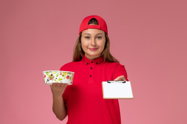 Corriere femminile di vista frontale in uniforme rossa e mantello che tiene il blocco note e la ciotola di consegna sul lavoratore di servizio di consegna di lavoro uniforme del fondo rosa