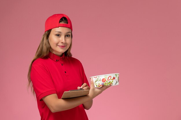 Corriere femminile di vista frontale in uniforme rossa e mantello che tiene il blocco note e la ciotola di consegna sul lavoro di servizio di consegna di lavoro uniforme del fondo rosa