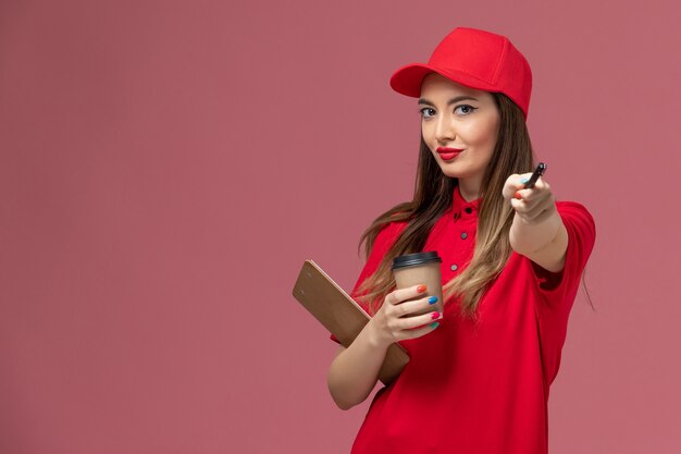 Corriere femminile di vista frontale in uniforme rossa e mantello che tiene il blocco note e la penna della tazza di caffè di consegna su fondo rosa lavoro uniforme di consegna di servizio del lavoratore