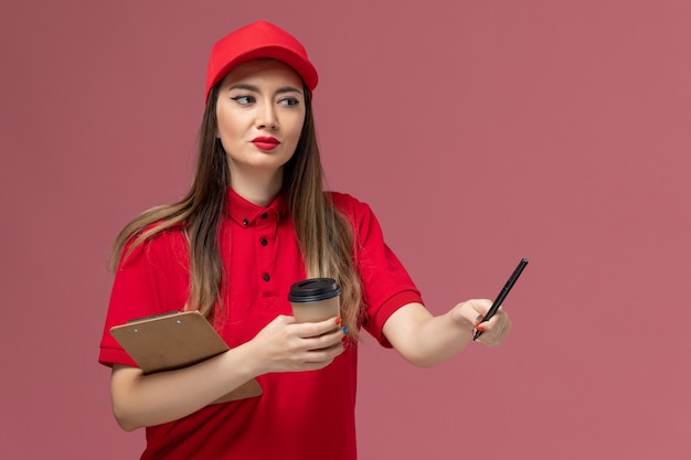 Corriere femminile di vista frontale in uniforme rossa e mantello che tiene il blocco note e la penna della tazza di caffè di consegna sullo sfondo rosa dell'uniforme di consegna del servizio
