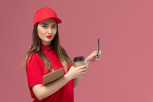 Corriere femminile di vista frontale in uniforme rossa e mantello che tiene il blocco note e la penna della tazza di caffè di consegna sullo sfondo rosa dell'uniforme di consegna del servizio