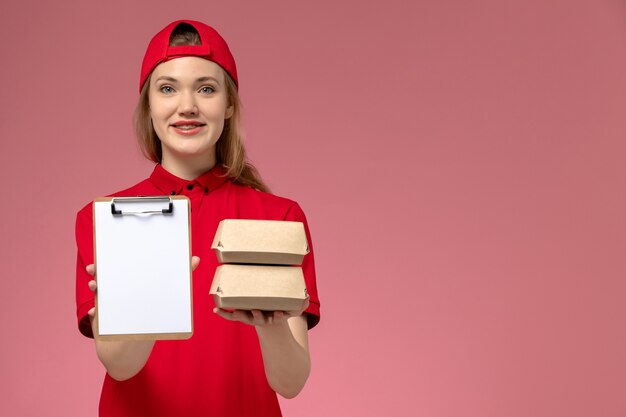 Corriere femminile di vista frontale in uniforme rossa e mantello che tiene il blocco note e piccoli pacchi di cibo di consegna che sorride sulla parete rosa chiaro, consegna uniforme di lavoro di servizio