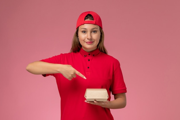 Corriere femminile di vista frontale in uniforme rossa e mantello che tiene il pacchetto di cibo di consegna sul lavoro dell'azienda di servizio di consegna uniforme sfondo rosa chiaro