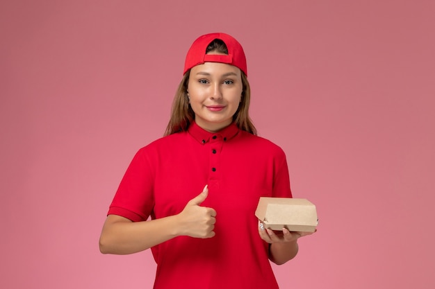 Corriere femminile di vista frontale in uniforme rossa e mantello che tiene il pacchetto di cibo per la consegna e sorridente sul muro rosa, lavoro di società di servizi di consegna uniforme