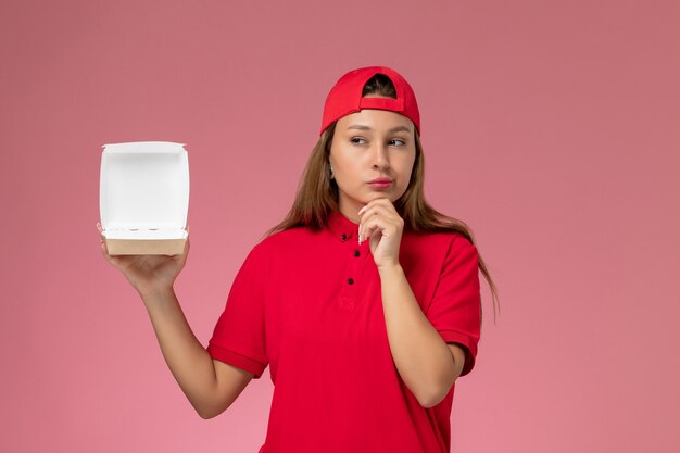 Corriere femminile di vista frontale in uniforme rossa e mantello che tiene il pacchetto di cibo vuoto di consegna e pensa sul muro rosa chiaro, società di servizi di consegna uniforme