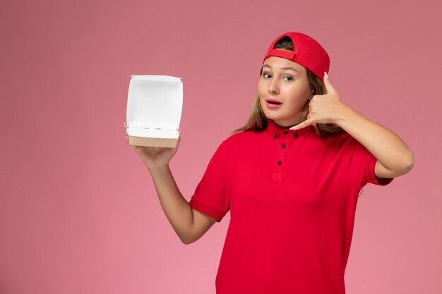 Corriere femminile di vista frontale in uniforme rossa e mantello che tiene il pacchetto di cibo vuoto di consegna sulla parete rosa chiaro, lavoratore di società di servizio di consegna uniforme