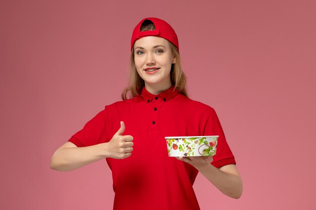 Corriere femminile di vista frontale in uniforme rossa e mantello che tiene la ciotola di consegna sulla parete rosa chiaro, lavoratore di consegna dell'uniforme di servizio