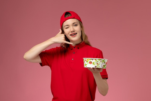 Corriere femminile di vista frontale in uniforme rossa e mantello che tiene la ciotola di consegna sulla parete rosa chiaro, lavoro dell'operaio di consegna dell'uniforme di servizio
