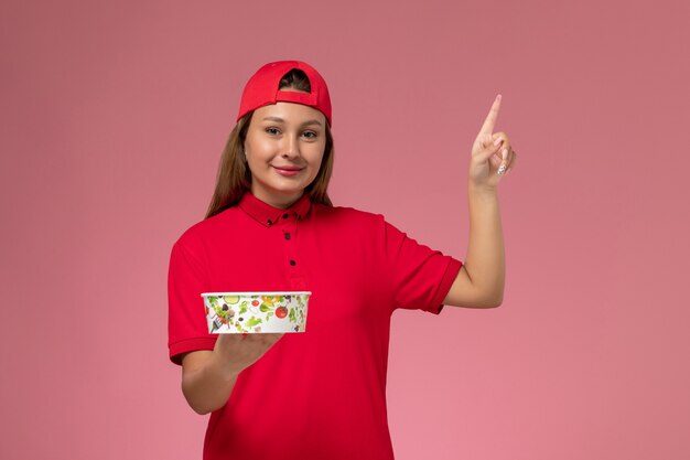 Corriere femminile di vista frontale in uniforme rossa e mantello che tiene la ciotola di consegna sulla parete rosa, lavoratore di lavoro di servizio di consegna uniforme