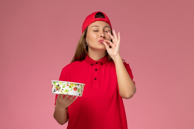 Corriere femminile di vista frontale in uniforme rossa e mantello che tiene la ciotola di consegna sulla parete rosa, ragazza di lavoro di servizio di consegna uniforme