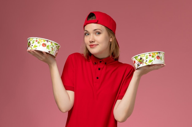 Corriere femminile di vista frontale in uniforme rossa e mantello che tiene le ciotole di consegna con un leggero sorriso sul muro rosa chiaro, consegna uniforme di servizio