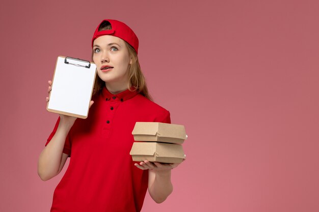 Corriere femminile di vista frontale in uniforme rossa e mantello che tiene piccoli pacchi di cibo di consegna pensando sulla parete rosa chiaro, consegna uniforme di servizio