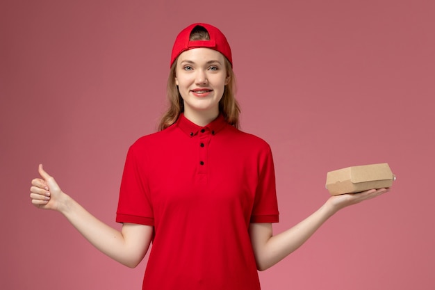 Corriere femminile di vista frontale in uniforme rossa e mantello che tiene piccolo pacchetto di cibo di consegna che sorride sulla parete rosa-chiaro, lavoro uniforme dell'azienda di servizio di consegna