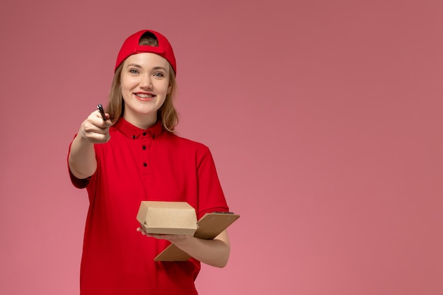 Corriere femminile di vista frontale in uniforme rossa e mantello che tiene piccolo pacchetto di cibo di consegna con blocco note e penna sul muro rosa, lavoratore di lavoro di consegna uniforme di servizio