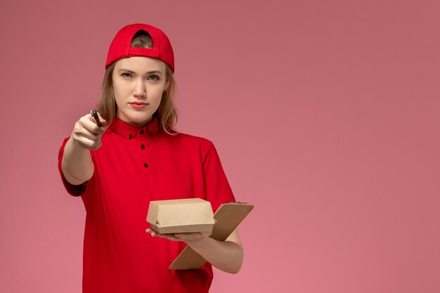 Corriere femminile di vista frontale in uniforme rossa e mantello che tiene piccolo pacchetto di cibo di consegna con blocco note e penna sul muro rosa, lavoro di consegna uniforme di servizio