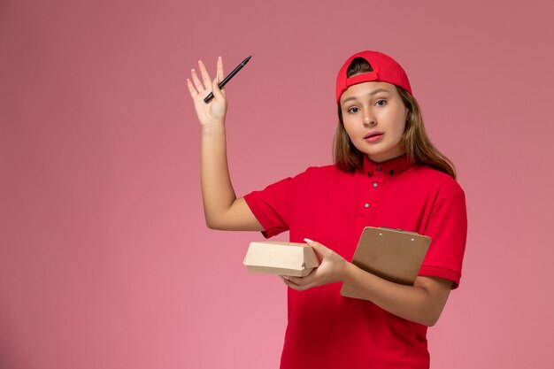 Corriere femminile di vista frontale in uniforme rossa e mantello che tiene piccolo pacchetto di cibo di consegna e blocco note sul muro rosa, lavoro di servizio di consegna del lavoratore uniforme