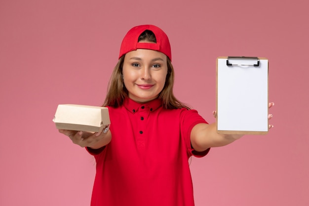 Corriere femminile di vista frontale in uniforme rossa e mantello che tiene piccolo pacchetto di cibo di consegna e blocco note sul muro rosa, società di lavoratori di servizio di consegna uniforme