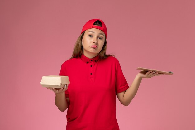 Corriere femminile di vista frontale in uniforme rossa e mantello che tiene piccolo pacchetto di cibo di consegna e blocco note sulla parete rosa, società di lavoro di servizio di consegna uniforme