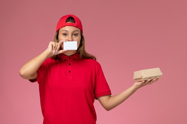 Corriere femminile di vista frontale in uniforme rossa e mantello che tiene piccolo pacchetto di cibo di consegna e carta sul muro rosa chiaro, servizio di consegna del lavoratore di lavoro uniforme