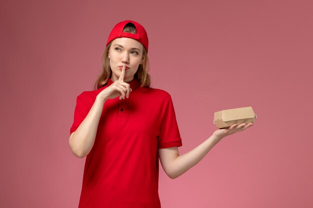 Corriere femminile di vista frontale in uniforme rossa e mantello che tiene piccolo pacchetto di cibo di consegna sulla parete rosa chiaro, lavoro uniforme di lavoro dell'azienda di servizio di consegna