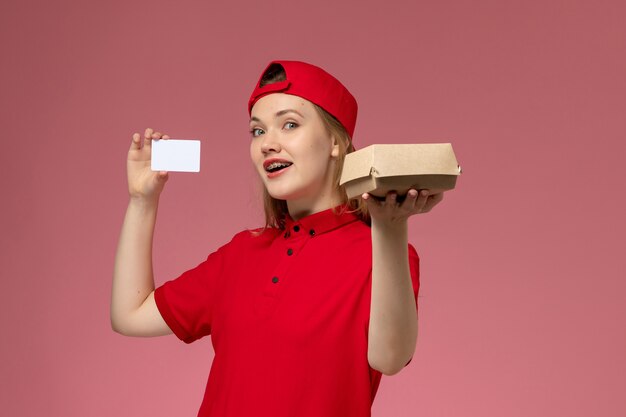 Corriere femminile di vista frontale in uniforme rossa e mantello che tiene piccolo pacchetto di cibo per la consegna con carta di plastica bianca sul muro rosa chiaro, consegna uniforme di lavoro