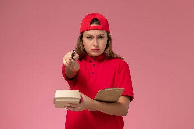 Corriere femminile di vista frontale in uniforme rossa e mantello che tiene piccolo pacchetto di cibo per la consegna e blocco note per scrivere note sul muro rosa, servizio di consegna uniforme