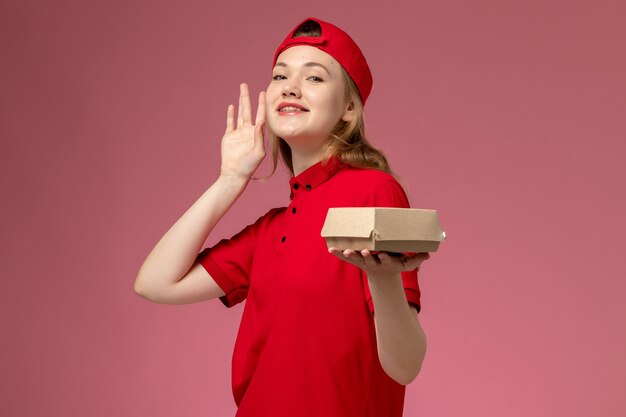 Corriere femminile di vista frontale in uniforme rossa e mantello che tiene piccolo pacchetto di cibo per la consegna sulla parete rosa chiaro, lavoro di lavoro uniforme della società di servizi di consegna