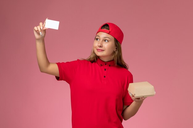 Corriere femminile di vista frontale in uniforme rossa e mantello che tiene poco pacchetto di cibo di consegna e carta sulla parete rosa, lavoro di lavoro di servizio di consegna uniforme