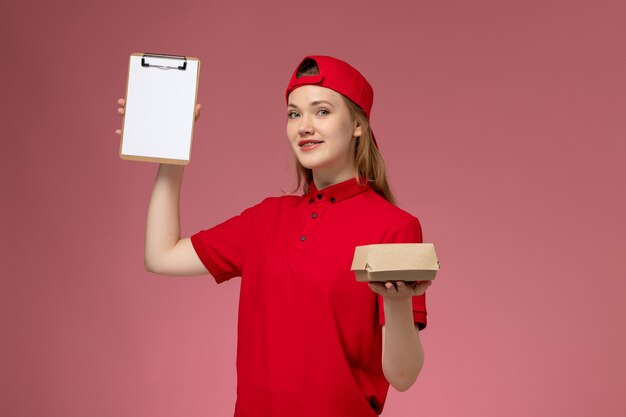 Corriere femminile di vista frontale in uniforme rossa e mantello che tiene un piccolo pacchetto di cibo per la consegna con il blocco note sul muro rosa, uniforme da lavoro dell'azienda di servizi di consegna