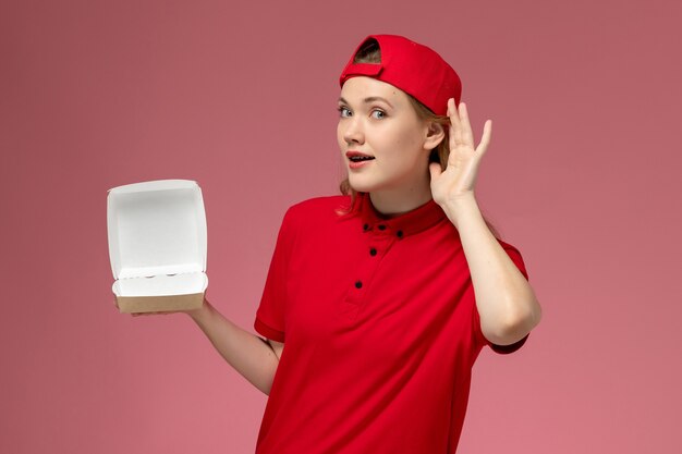 Corriere femminile di vista frontale in uniforme rossa e mantello che tiene un piccolo pacchetto di cibo vuoto di consegna cercando di ascoltare sul muro rosa, lavoro uniforme della società di servizi di consegna