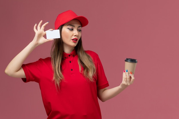 Corriere femminile di vista frontale in uniforme rossa e tazza di caffè di consegna della tenuta del capo con carta bianca sull'uniforme di consegna di lavoro di servizio del fondo rosa