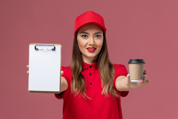 Corriere femminile di vista frontale in uniforme rossa e tazza di caffè di consegna della tenuta del capo con il blocco note sull'uniforme di consegna di lavoro di servizio del fondo rosa chiaro
