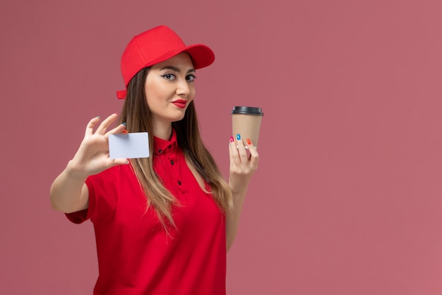 Corriere femminile di vista frontale in uniforme rossa e tazza di caffè di consegna della tenuta del capo con la carta bianca che sorride sull'uniforme rosa dell'operaio di consegna di lavoro di servizio del fondo