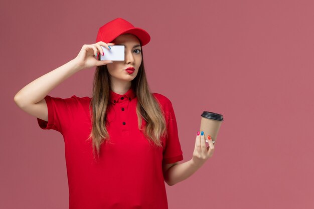 Corriere femminile di vista frontale in uniforme rossa e tazza di caffè di consegna della tenuta del capo con la carta bianca sull'uniforme rosa di consegna del servizio di scrivania