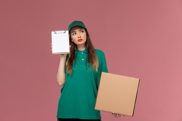 Corriere femminile di vista frontale in uniforme verde che tiene il blocco note della scatola dell'alimento e che pensa sul lavoro di consegna dell'uniforme del lavoratore di servizio dell'azienda della parete rosa