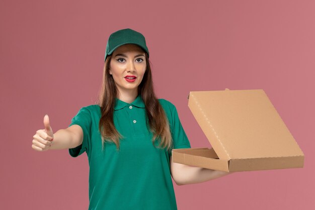 Corriere femminile di vista frontale in uniforme verde che tiene la scatola di consegna del cibo e che posa sulla consegna dell'uniforme di servizio dell'azienda della parete rosa