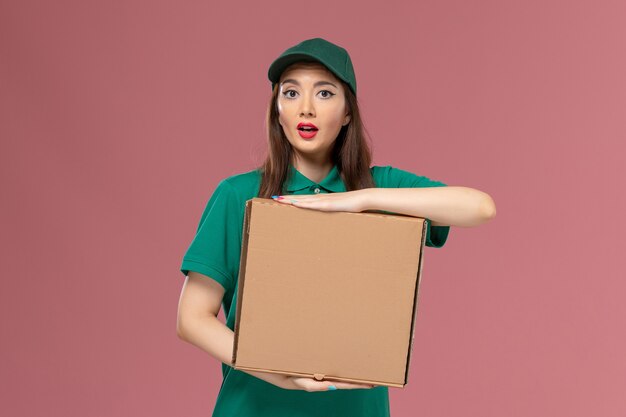 Corriere femminile di vista frontale in uniforme verde che tiene la scatola di consegna del cibo sulla parete rosa lavoro lavoratore servizio uniforme ragazza consegna azienda