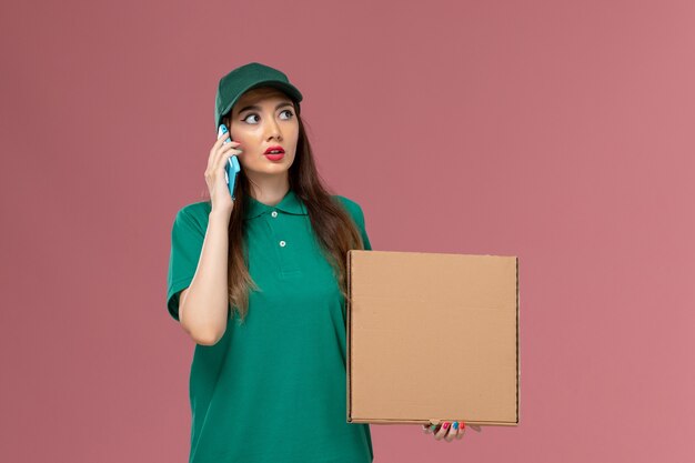 Corriere femminile di vista frontale in uniforme verde che tiene la scatola di consegna del cibo usando il suo telefono sul lavoro di consegna dell'uniforme di servizio dell'azienda della parete rosa