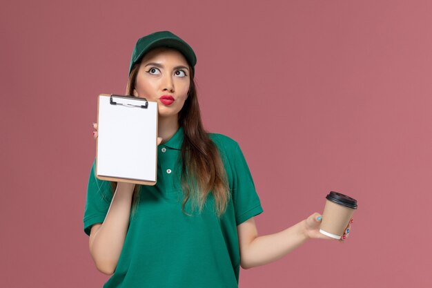 Corriere femminile di vista frontale in uniforme verde e capo che tiene la tazza di caffè di consegna e il blocchetto per appunti che pensa sulla femmina dell'operaio del lavoro di consegna dell'uniforme di lavoro di servizio della parete rosa