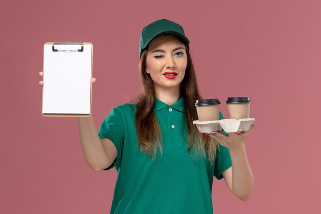 Corriere femminile di vista frontale in uniforme verde e mantello che tiene le tazze di caffè di consegna e blocchetto per appunti sulla consegna uniforme dell'operaio di lavoro di servizio della parete rosa