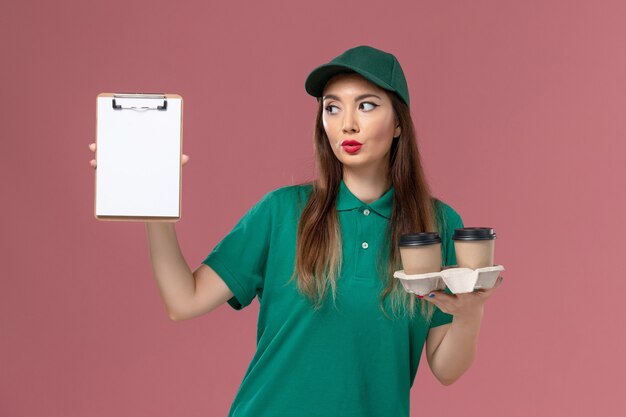 Corriere femminile di vista frontale in uniforme verde e mantello che tiene le tazze di caffè di consegna e blocco note che posano sulla consegna dell'uniforme di lavoro di servizio della parete rosa