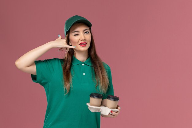 Corriere femminile di vista frontale in uniforme verde e mantello che tiene le tazze di caffè di consegna sul muro rosa azienda servizio lavoro uniforme consegna operaio femminile lavoro