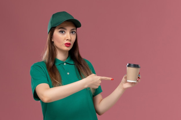 Corriere femminile di vista frontale in uniforme verde e tazza di caffè di consegna della tenuta del capo che sorride sul lavoro di consegna dell'uniforme di lavoro di servizio della parete rosa