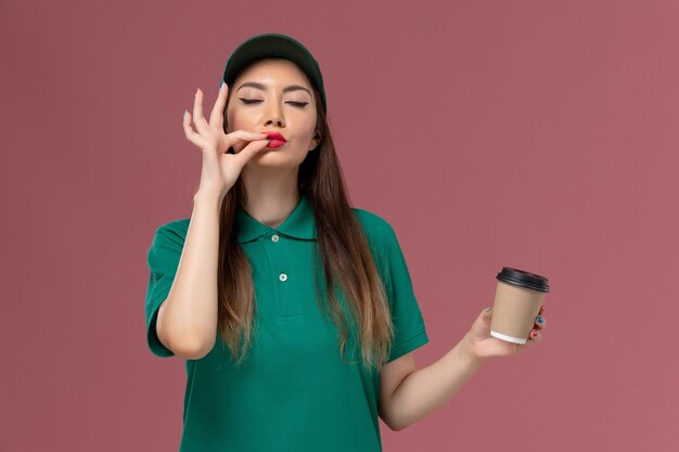 Corriere femminile di vista frontale in uniforme verde e tazza di caffè di consegna della tenuta del capo che sorride sul lavoro di consegna dell'uniforme di lavoro di servizio della scrivania rosa