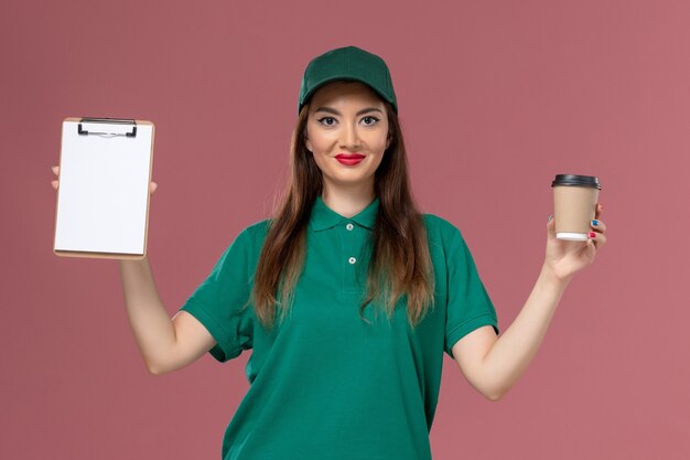 Corriere femminile di vista frontale in uniforme verde e tazza di caffè di consegna della tenuta del capo e blocchetto per appunti sulla società di lavoro di consegna dell'uniforme di lavoro di servizio della parete rosa