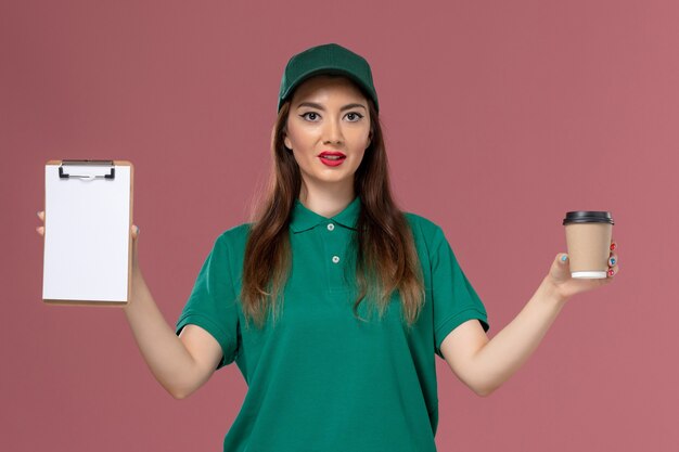 Corriere femminile di vista frontale in uniforme verde e tazza di caffè di consegna della tenuta del capo e blocco note sulla società di consegna dell'uniforme di lavoro di servizio della parete rosa