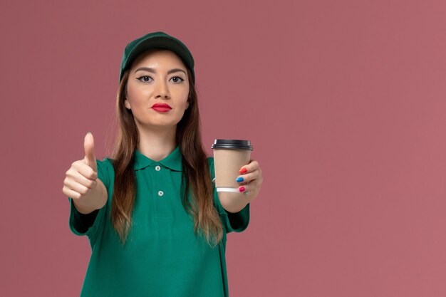 Corriere femminile di vista frontale in uniforme verde e tazza di caffè di consegna della tenuta del capo sulla consegna uniforme di lavoro di servizio della parete rosa chiaro