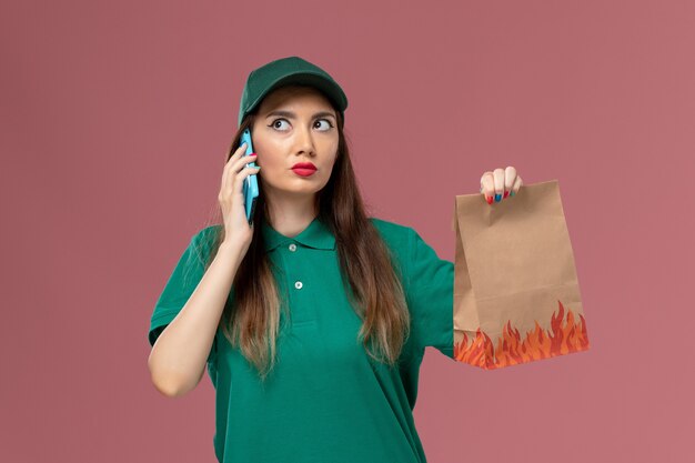 Corriere femminile di vista frontale in uniforme verde, parlando al telefono e tenendo il pacchetto di cibo sul lavoro di consegna uniforme di servizio parete rosa